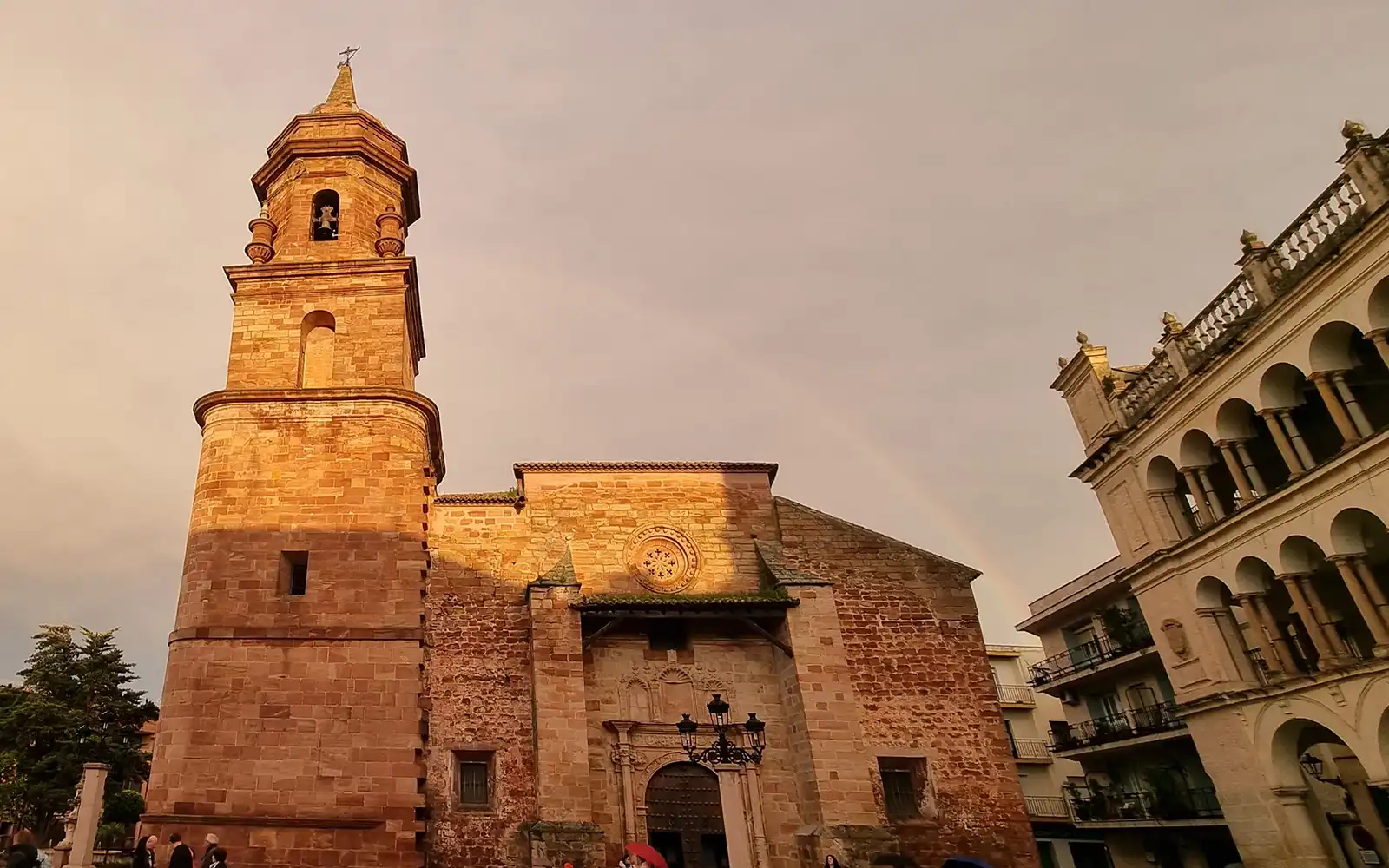 Iglesia de piedra con torre campanario y espadaña en la plaza del municipio, flanqueada por edificios con galería de arcos, bajo luz de tarde
